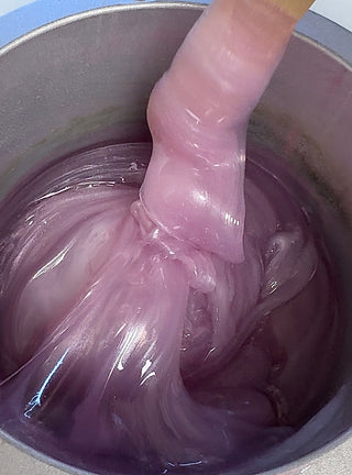A close-up view of a waxing strip being lifted out of a container filled with purple hard wax, illustrating the texture and consistency of the product.